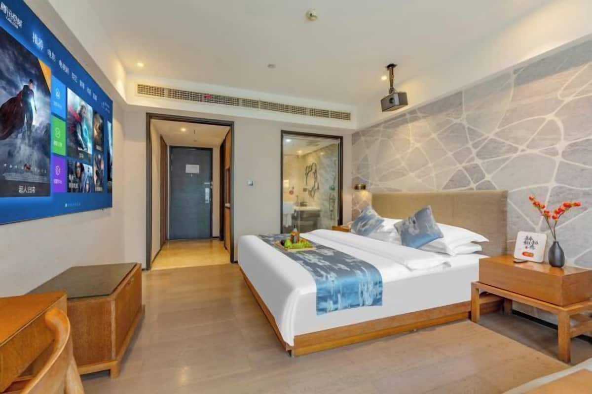 A modern hotel room with a large bed, a wooden nightstand, a desk, and a wall-mounted TV displaying movie posters.