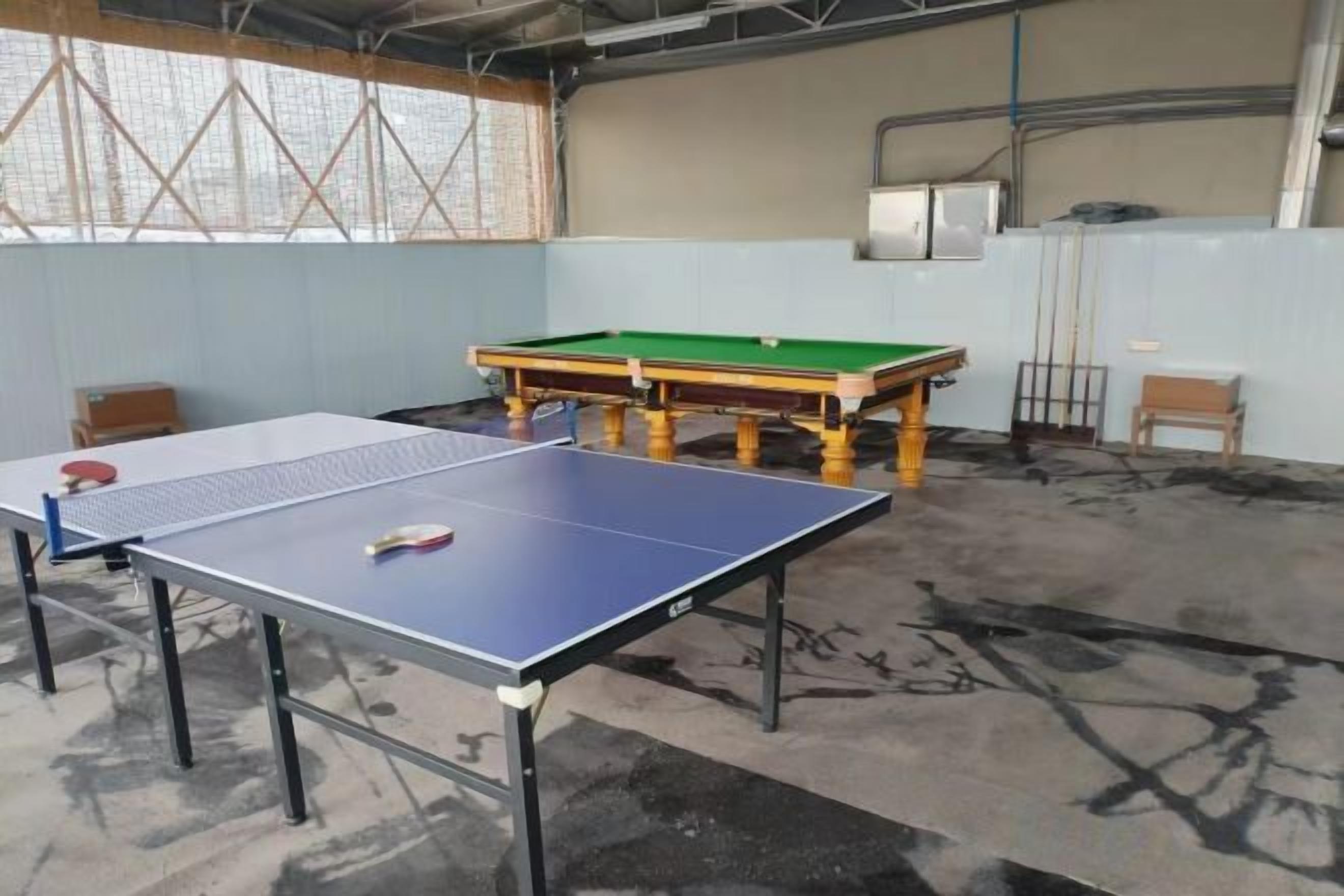 A ping pong table and a billiards table in a room with concrete flooring and a high ceiling.