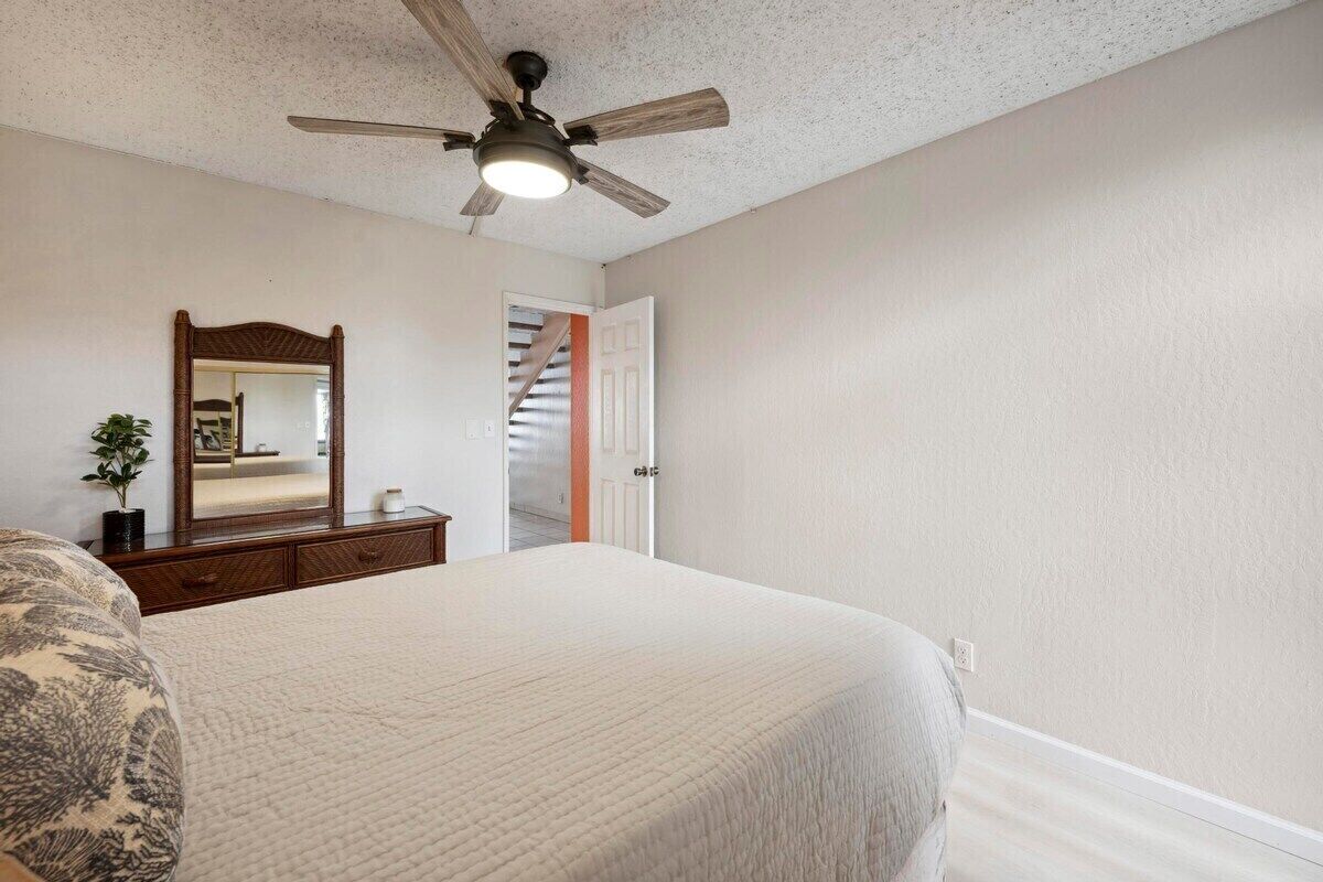 A bedroom with a bed, a ceiling fan, a dresser, a mirror, and a door leading to a staircase.