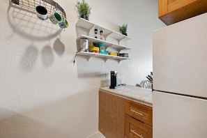 Una cocina con paredes blancas, armarios de madera y encimera blanca. Hay tres repisas blancas con diversos objetos, incluyendo una cafetera, una caja y una planta.