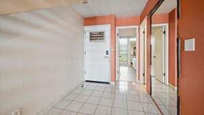 A hallway with white tiled floors, a white door with a window, and a door leading to a room with a bed. - Oceanfront Maui Condo-Walk to Beach, Shops, Cafés (Lahaina)