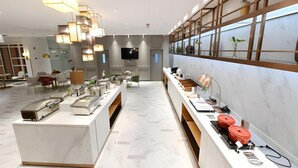 A modern hotel lobby with a central buffet area, a flat-screen TV, and a large window with plants. - Magnotel (Qinzhou)