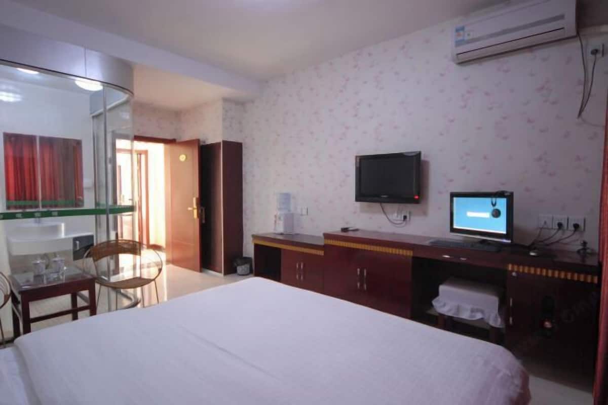 A hotel room with a bed, a desk with a computer, a TV, and a glass partition separating the room from the hallway.