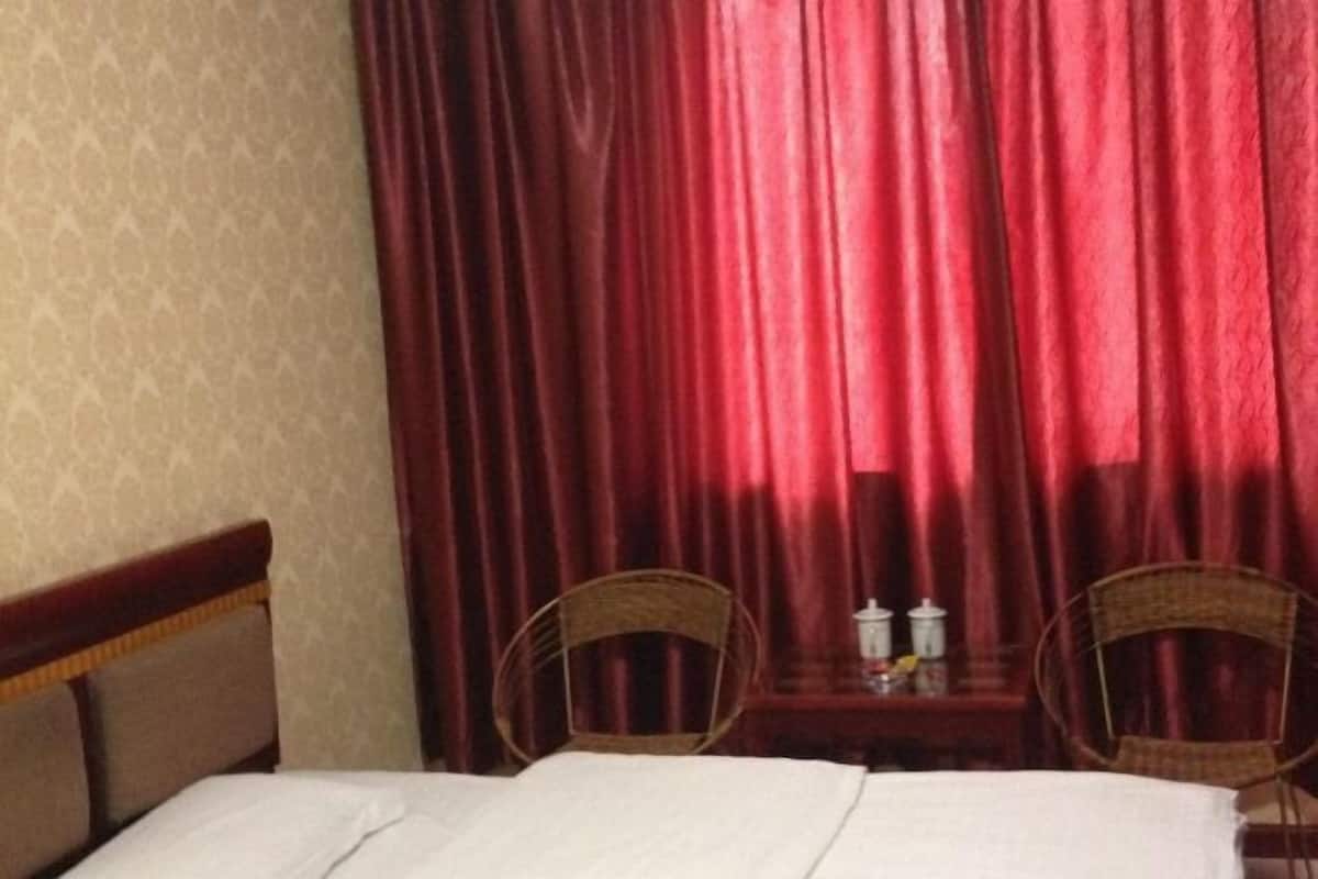 A hotel room with a bed, two wicker chairs, a small table with a lamp, and red curtains.
