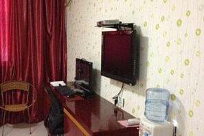 Quarto com escrivaninha, cadeira, dispensador de água e televisão na parede.