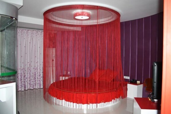 Uma cama redonda com dossel vermelho, uma televisão sobre suporte e uma parede roxa.