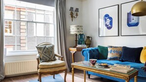A living room with a blue sofa, a patterned armchair, a glass coffee table, and a window with a view of a brick building.