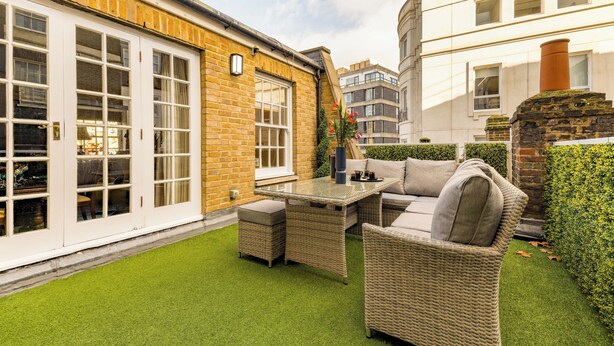 A patio with wicker furniture and a glass table, surrounded by a well-maintained lawn and a brick wall.