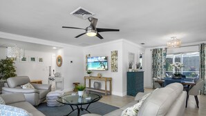 A modern living room with a ceiling fan, a flat-screen TV, a dining area with a table and chairs, and a large window with curtains. - Your vacation home away from home (Naples)