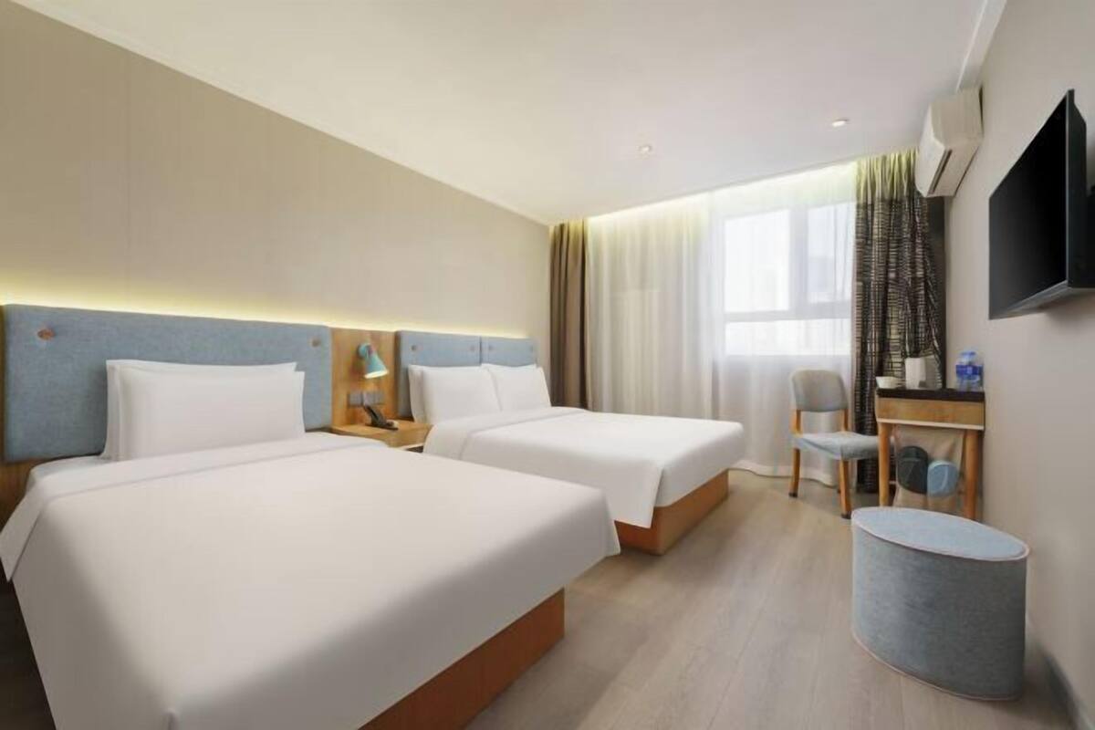 Quarto de hotel com duas camas, uma escrivaninha com cadeira, televisão e janela com cortinas.