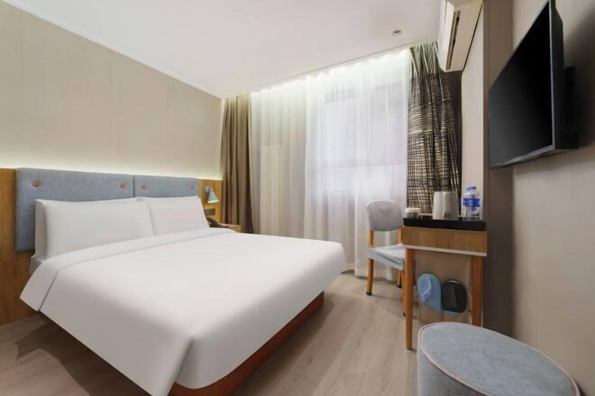 Quarto de hotel com cama, escrivaninha com cadeira, televisão e janela com cortinas.