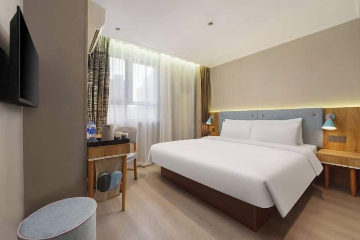 Quarto de hotel com uma cama grande, uma escrivaninha com cadeira, televisão na parede e mesa de cabeceira com luminária.