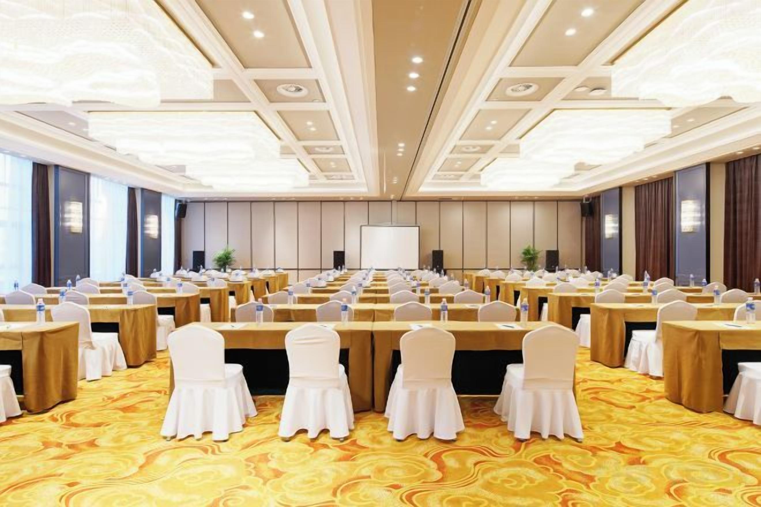 A conference room with rows of tables and chairs, a large screen at the front, and a carpeted floor.