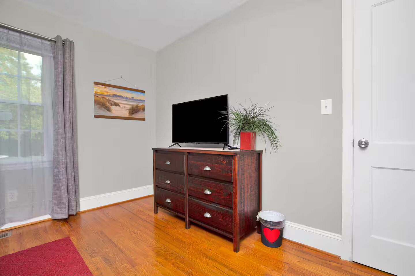 Una habitación con un aparador de madera, un televisor encima, una planta en maceta, un cuadro en la pared y un cubo de basura rojo con forma de corazón.