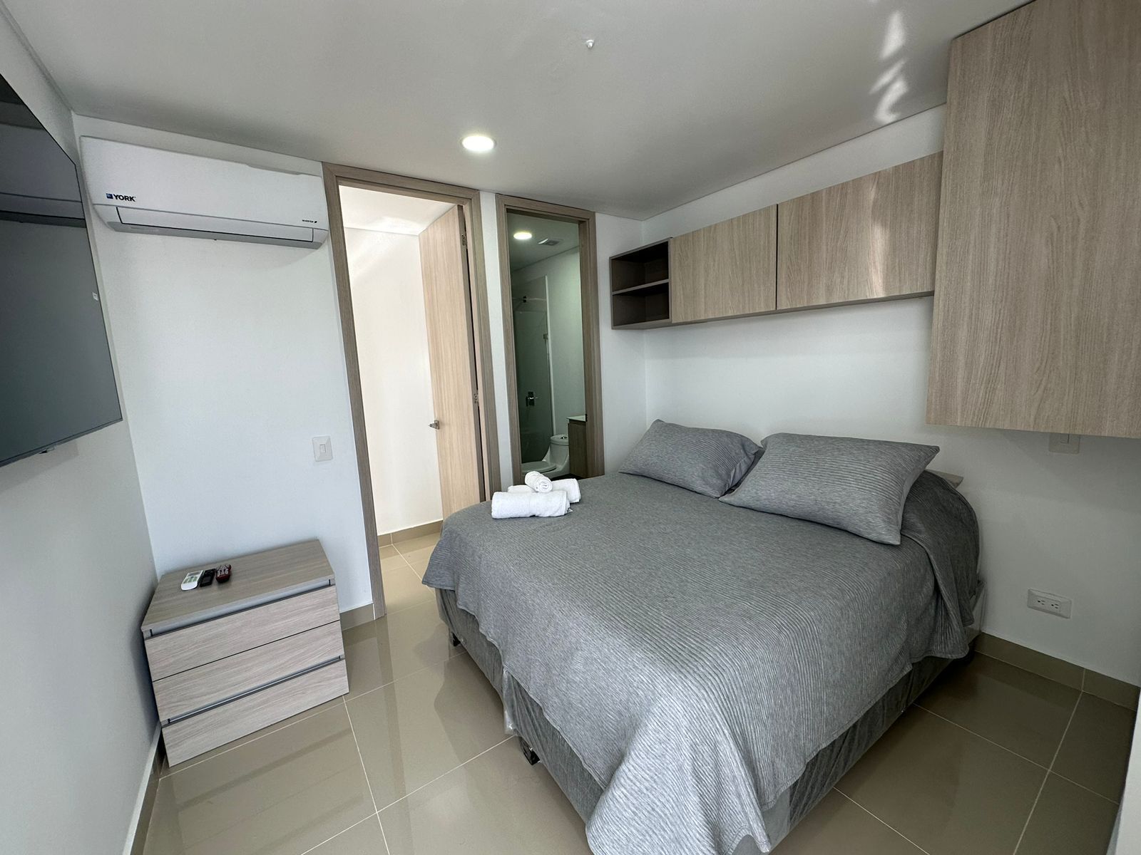 A modern bedroom with a bed, a wardrobe, a built-in TV, and a bathroom visible through an open door.