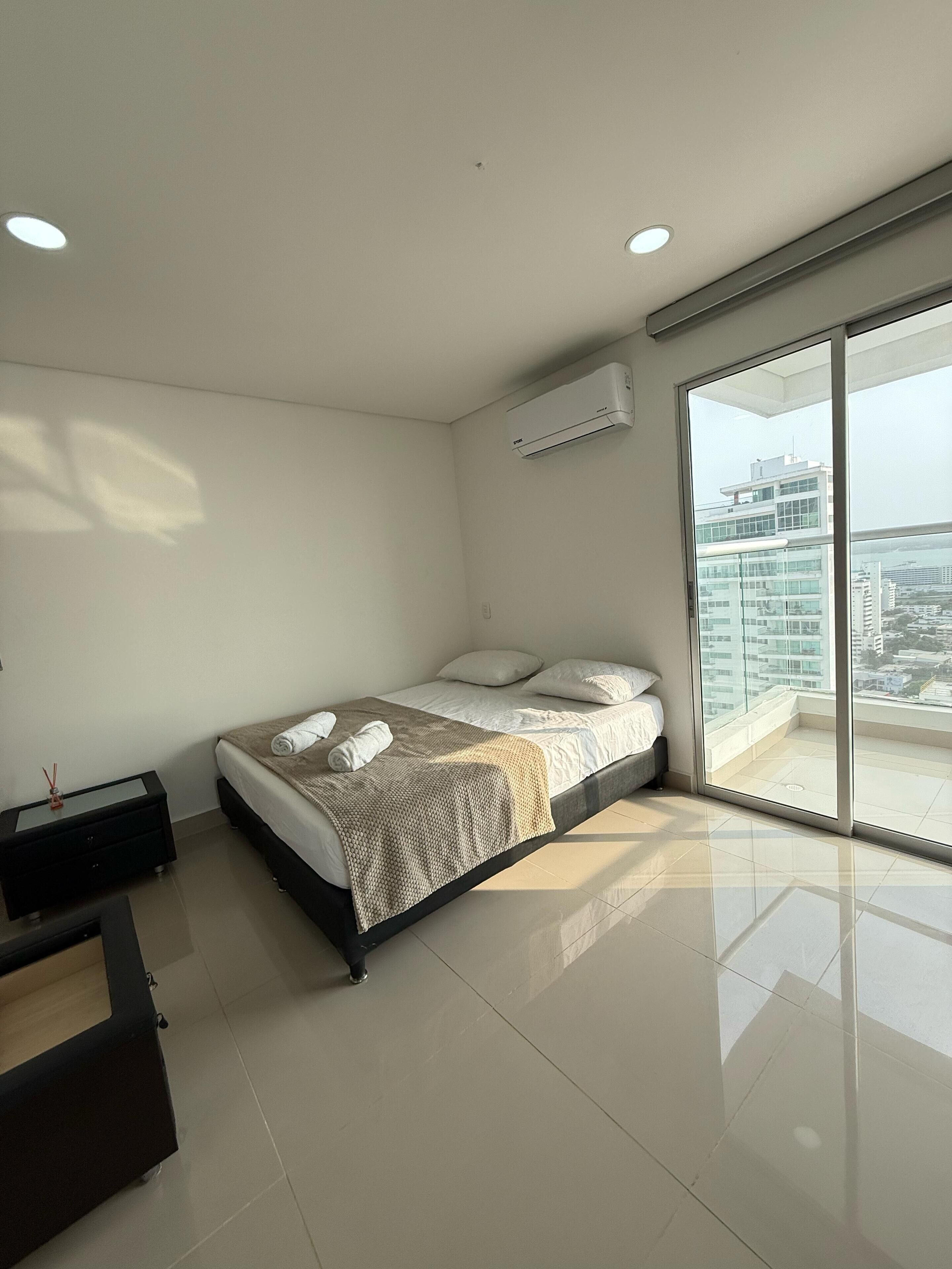 A modern bedroom with a bed, a bedside table, a built-in bench, a large window with a city view, and an air conditioning unit.