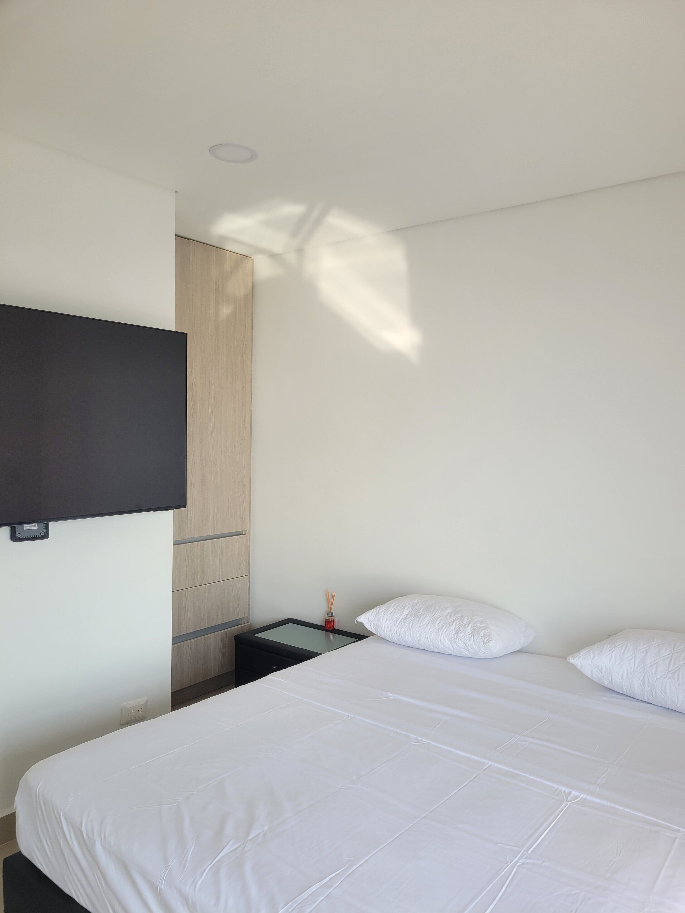 A modern hotel room with a large bed, a flat-screen TV mounted on the wall, a wooden wardrobe, and a small table with a red candle.