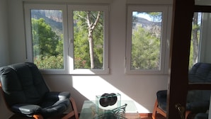 A room with three windows, a black leather chair, a glass table, and a view of a mountainous landscape. - Apartamento Deluxe (Oropesa del Mar)