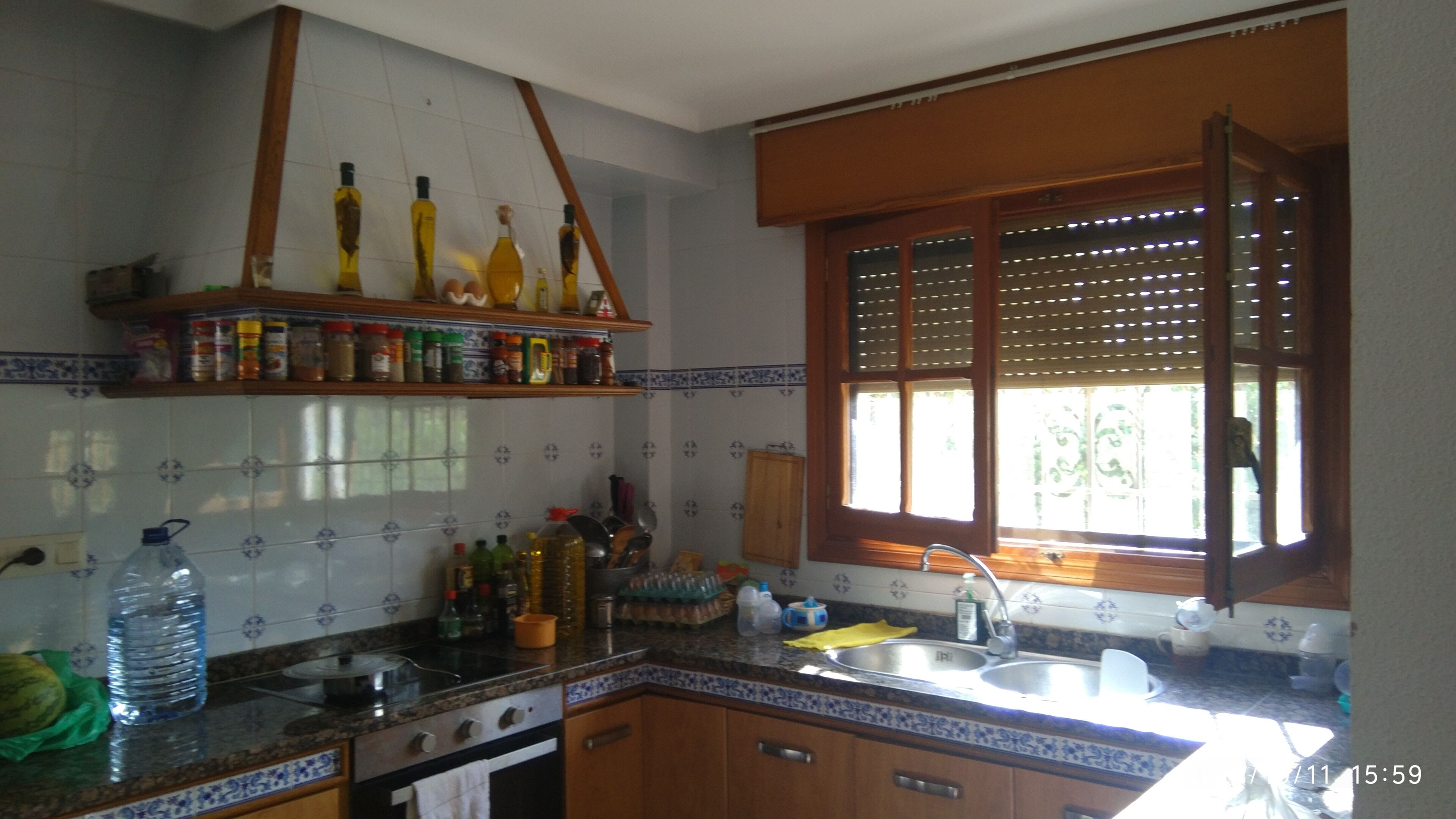 A kitchen with a tiled backsplash, a wooden countertop, a window with blinds, and a shelf with bottles.