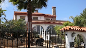 A large building with a tiled roof and a chimney, surrounded by a wrought iron fence and palm trees. - Apartamento Deluxe (Oropesa del Mar)