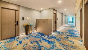 Un couloir d’hôtel avec un tapis à motifs, des portes en bois, un comptoir de réception et un couloir menant à une autre pièce.