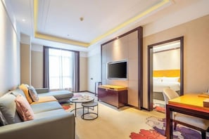 A modern hotel room with a sofa, a dining table, a TV, and a large window.
