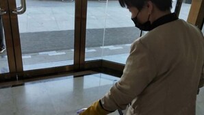 A person in a beige coat and yellow gloves standing in front of a glass door with a metal handle. - Shuiyi Jianianhua Hotel (Juancheng)