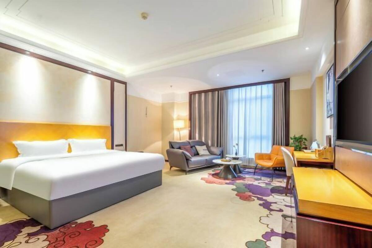A hotel room with a bed, a sofa, a desk, a TV, and a large window with curtains.