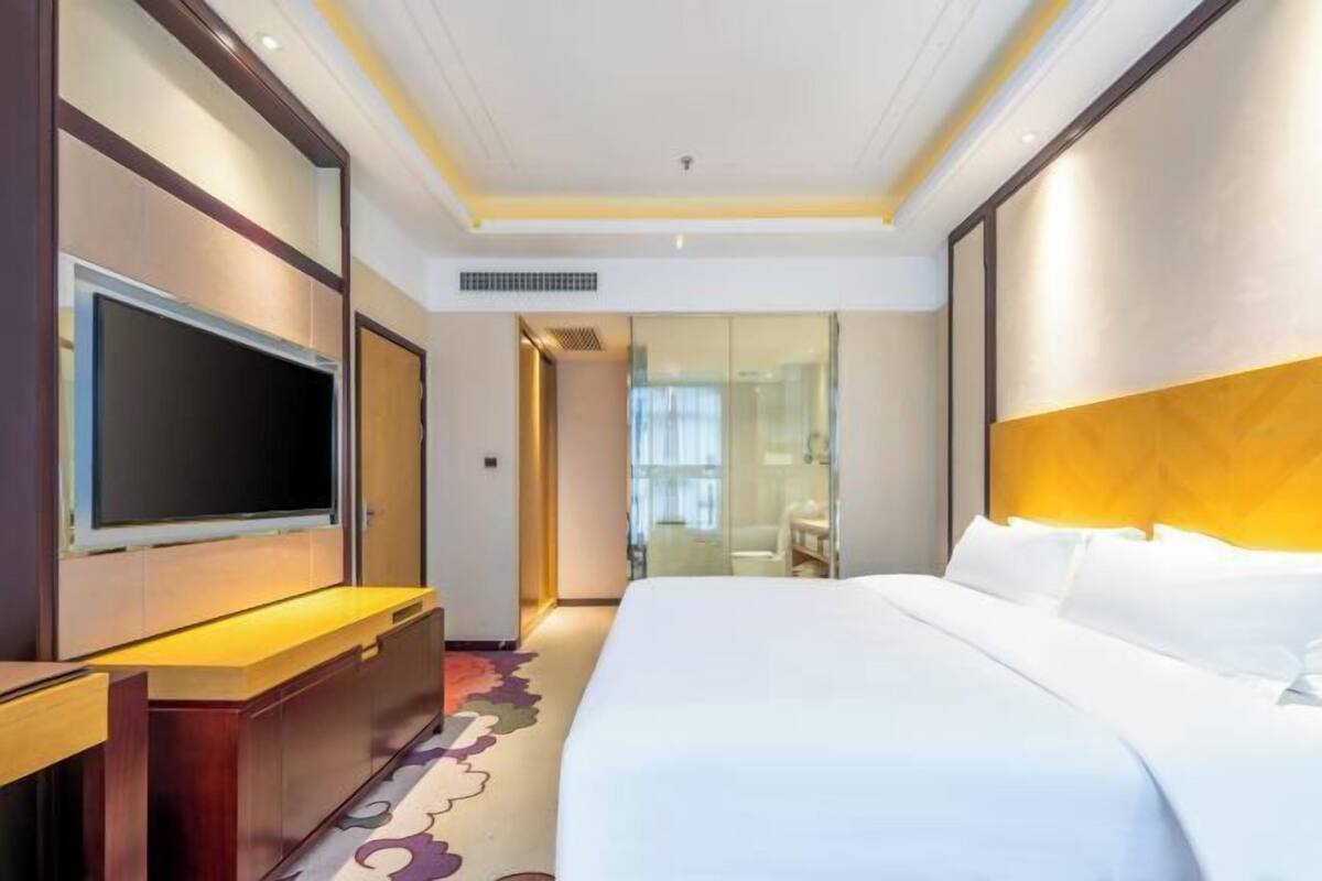 A hotel room with a large bed, a flat-screen TV mounted on the wall, a desk, and a bathroom visible through a glass door.