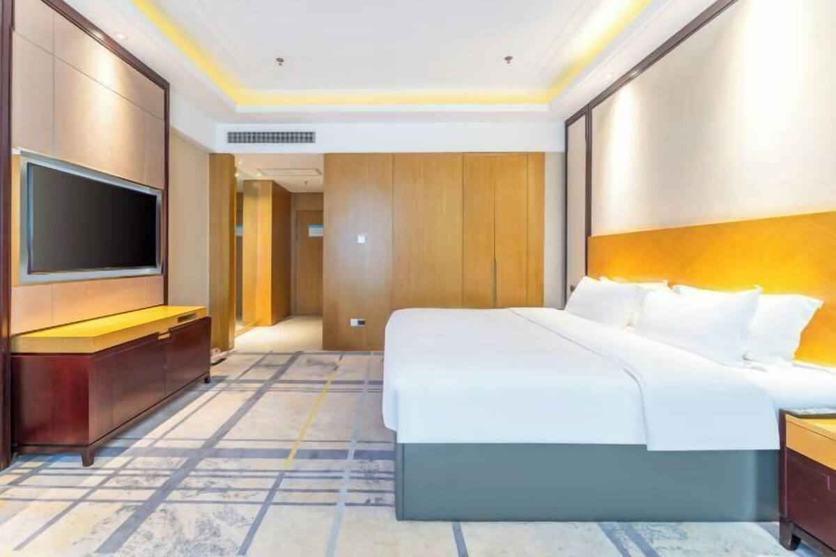 A hotel room with a bed, a TV, a wooden wardrobe, and a carpeted floor.
