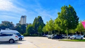 A parking lot with several cars, a white van, and a police car. There are trees and a building in the background. - Shuiyi Jianianhua Hotel (Juancheng)