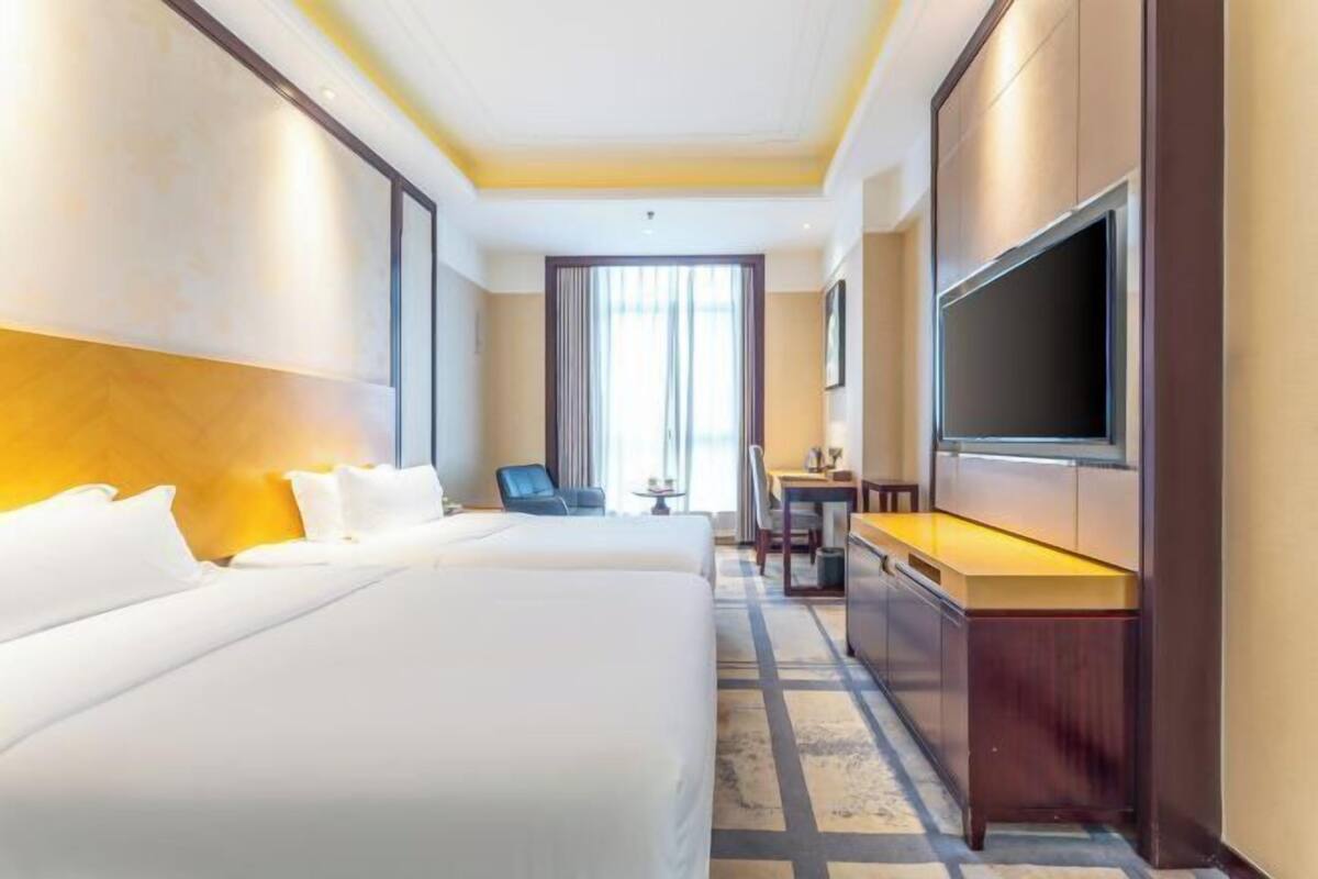 A hotel room with two beds, a desk, a chair, a TV, and a large window with curtains.