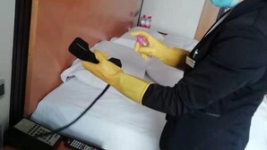 A person in a black jacket and yellow gloves cleaning a bed with a spray bottle and cloth. - Shuiyi Jianianhua Hotel (Juancheng)
