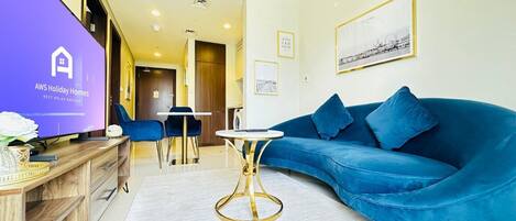 Un salon moderne avec un canapé bleu, une table ronde dorée et un téléviseur diffusant une publicité pour « AWS Holiday Homes ».