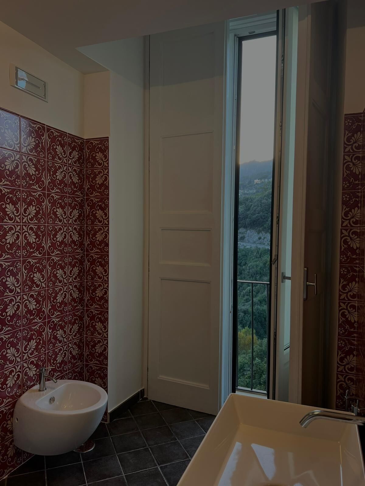 Casa de banho com lavatório branco, bidé e porta que dá para uma varanda com vista para o verde.