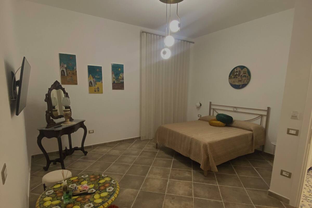 A small, well-lit room with a bed, a round table with chairs, and a TV mounted on the wall.