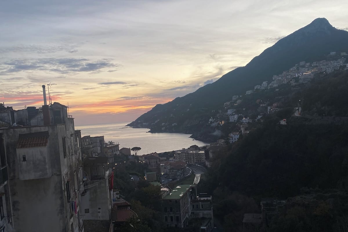 A coastal town with buildings along the hillside, a mountain in the background, and a sunset over the sea.
