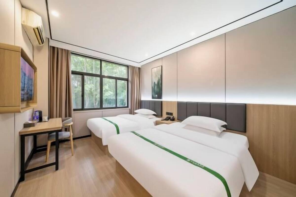 A hotel room with two beds, a desk, a chair, a nightstand, and a window with a view of trees.