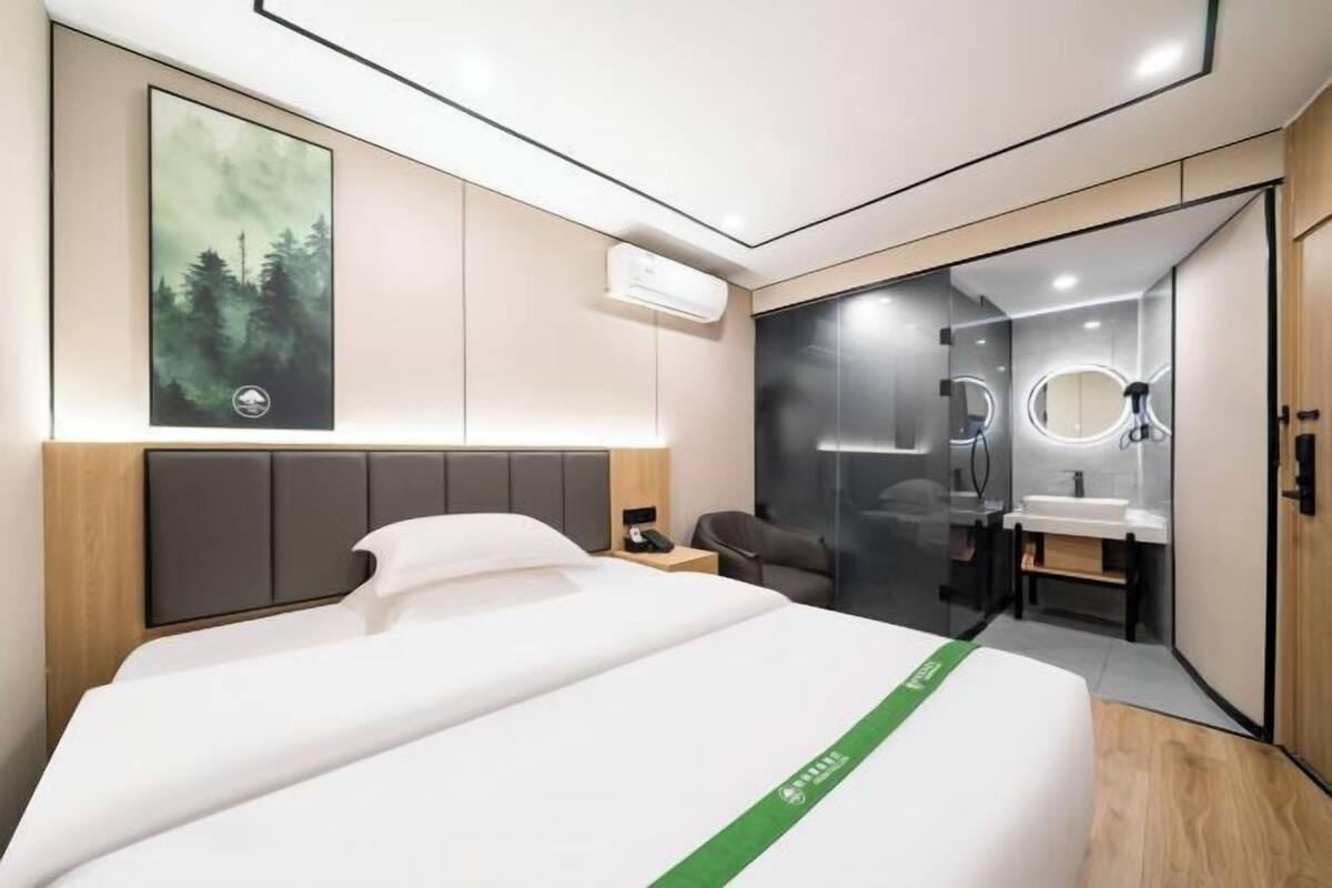 A modern hotel room with a bed, bedside table, chair, and a bathroom with a mirror and sink.
