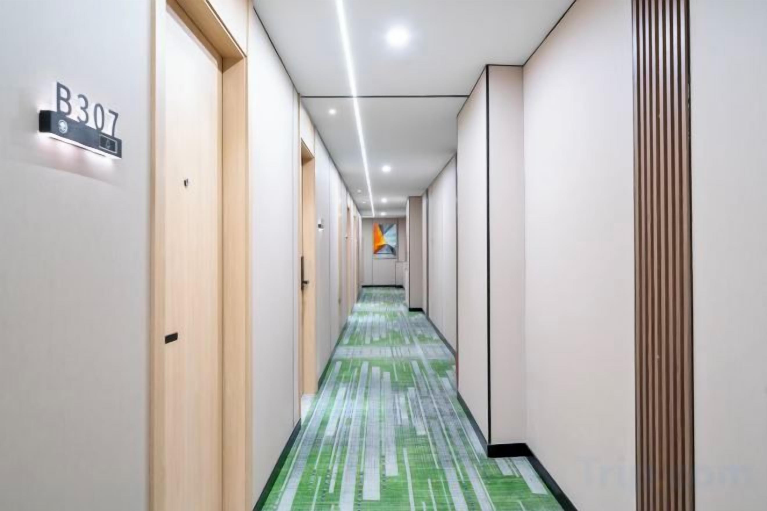 Un pasillo de hotel con alfombra de estampado verde, puertas numeradas y un diseño moderno en el techo.