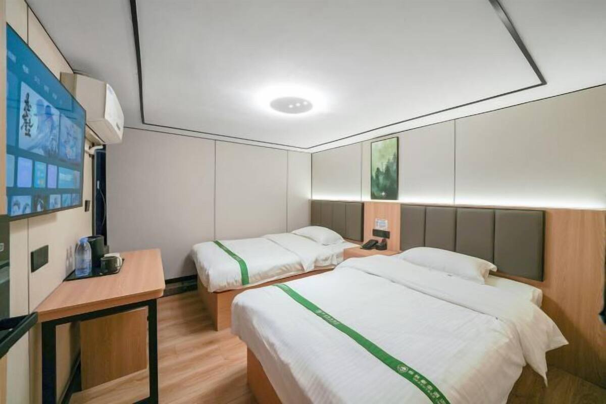 A hotel room with two beds, a desk, a chair, a small table, a wall-mounted TV, and a large window.