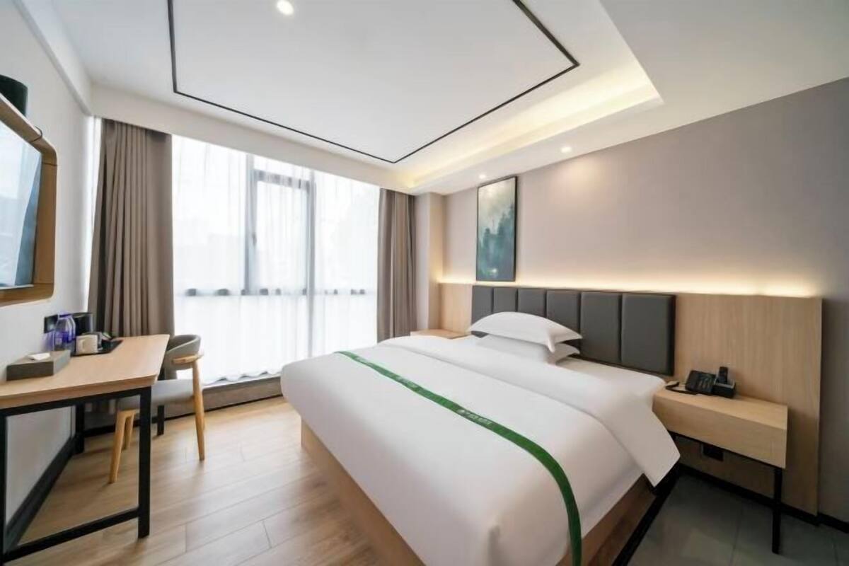 A hotel room with a bed, bedside table, desk, chair, and a large window.