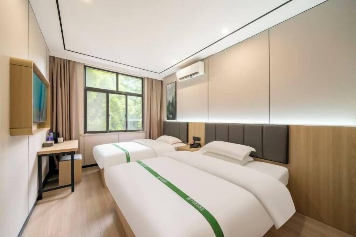 A hotel room with two beds, a desk, a TV, and a window with a view of trees.