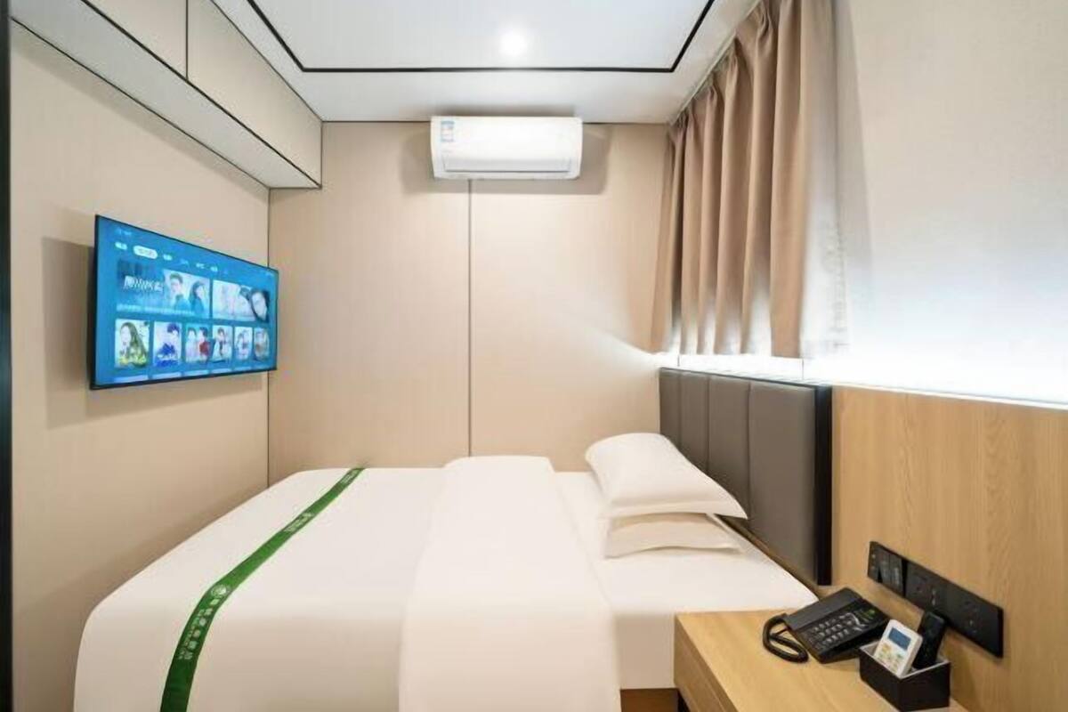A hotel room with a bed, a mounted television, an air conditioning unit, a wooden desk with a phone, and a window with curtains.