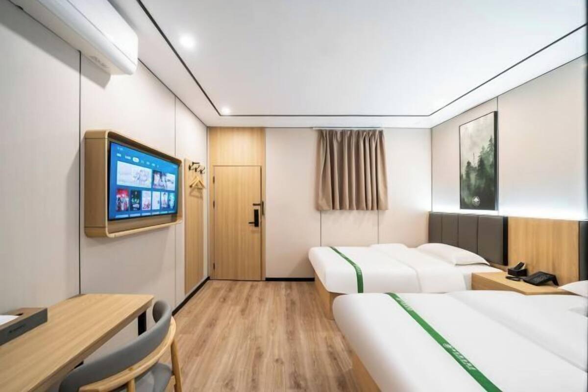 A hotel room with a wooden floor, a desk with a chair, two beds with green bedspreads, a mounted TV, a door, a window with curtains, and a framed picture on the wall.