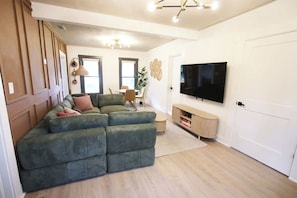 A living room with a sectional sofa, a flat-screen TV, a dining area, and a door.