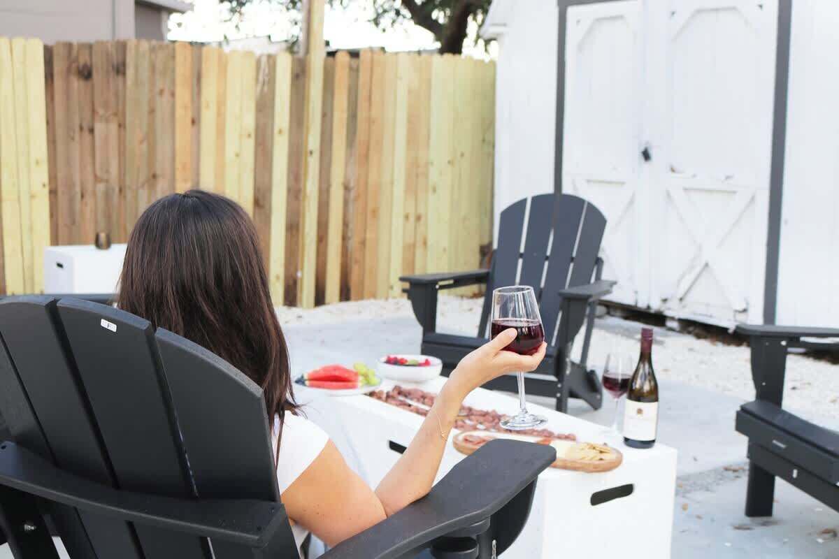 A person seated at an outdoor table with a wine glass, a bottle of wine, and food items.