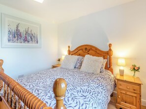A bedroom with a wooden bed frame, a patterned bedspread, a nightstand with a lamp, and a framed floral artwork on the wall.