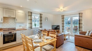 Une cuisine moderne avec des meubles blancs, un coin repas avec des meubles en bois, un salon avec un canapé et une télévision, et un balcon avec des sièges d’extérieur.