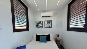 Een compacte hotelkamer met een bed, een airconditioningapparaat, twee kunstwerken aan de muur, een nachtkastje met een plant en een klein nachtkastje.